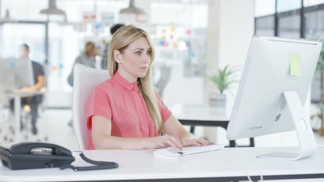 Close up young woman working on computer in creative office with colleagues talking in background
