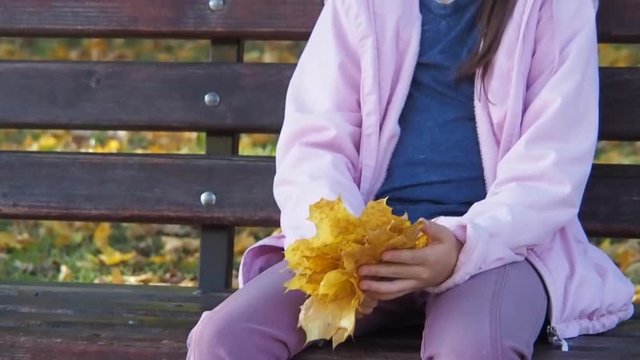 Sad child in an autumn park. Little girl with yellow leaves on a bench.