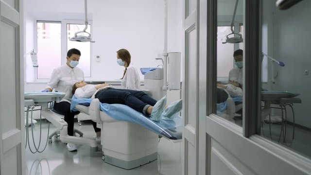 Patient sits down on blue dentist chair, assistant of dentist help to him.