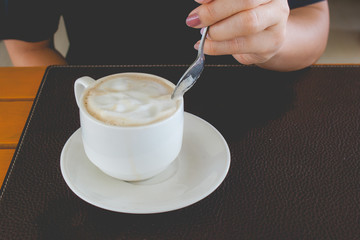 Woman hand holding coffee spoon and stirring hot coffee in white mug on wooden table at the cafe in vintage style.