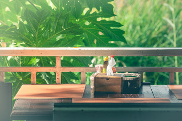 Wooden tissue box and garnish in basket (salt, pepper, toothpick, sauce) on table with green trees and sunlight background in the morning. (Selective focus)