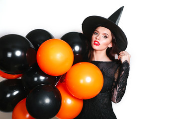 Portrait of beautiful young woman in black witch halloween costume with orange and black balloons over white background