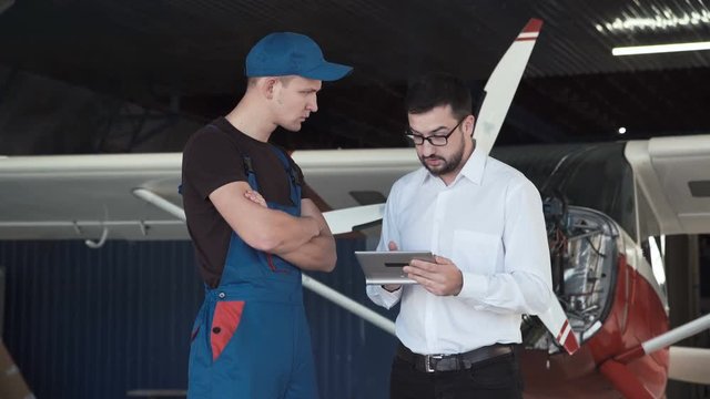 Mechanic and flight engineer having a discussion looking at a tablet-pc together as they stand in front of a small single engine aircraft in a hangar.