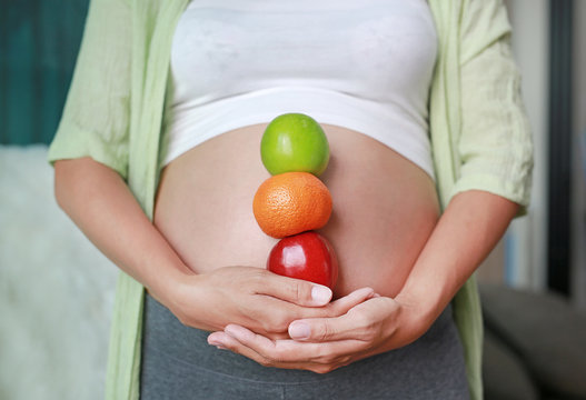 Pregnant woman holding Green-Red Apple and Orange fruit in vertical at her belly. Healthy Lifestyle.
