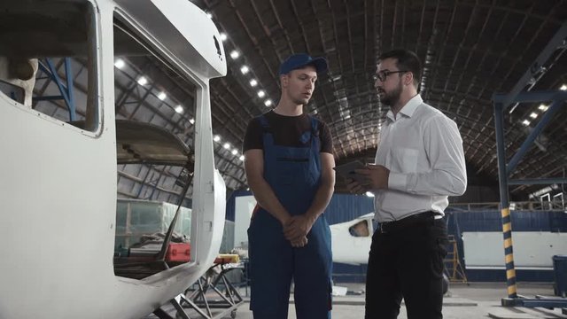 Aircraft mechanic and draftsman standing near plane carcass and talking with each other.