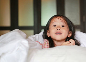 Happy little asian girl looking at camera with lying under blanket on the bed.