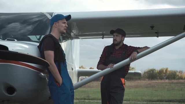 Pilot and mechanic chatting on an airfield as they stand together leaning on a small single engine plane