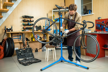 Obraz premium Young handsome mechanic wearing apron repairing bicycle while listening to music in headphones, interior of modern workshop on background