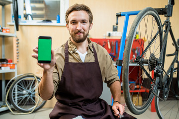 Portrait of smiling bearded mechanic sitting on haunches and showing smartphone with blank screen...
