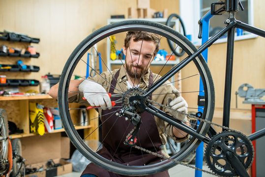 Enthusiastic young mechanic wearing apron sitting on haunches and adjusting bicycle chain, interior of modern workshop on background