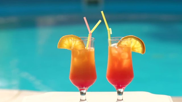 Cold refreshing drinks with fruits and straws are on the table near the pool. Two orange cocktail on a background of the pool. Slow motion.