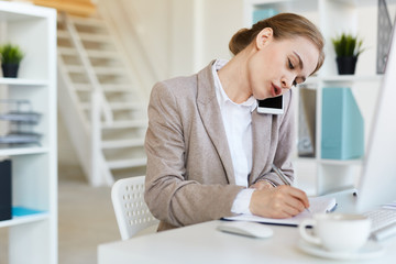 Busy secretary making notes in notebook while talking by smartphone in office