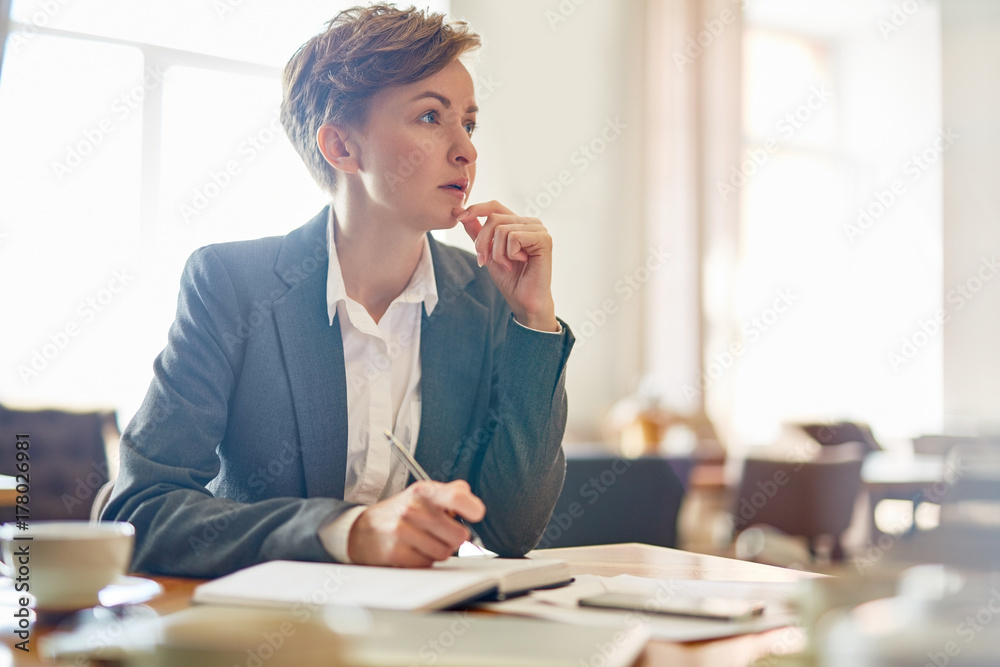 Wall mural Waist-up portrait of confident businesswoman taking necessary notes while thinking over promising project, interior of modern coffeehouse on background