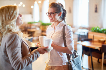 Profile view of joyful young manager holding paper cups of cappuccino in hand while standing at counter and chatting with female colleague, interior of cozy coffeehouse on background