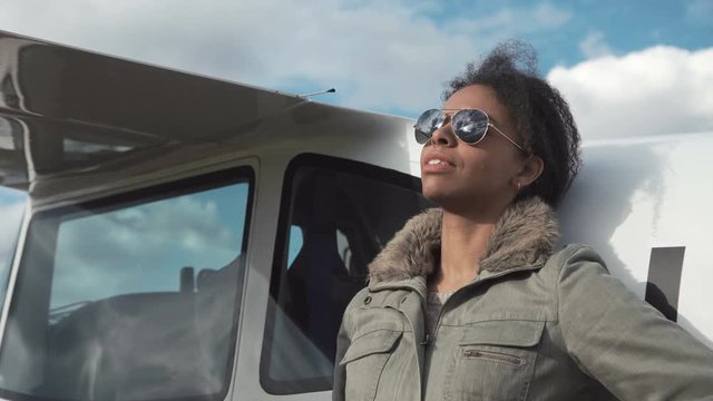 Attractive woman pilot wearing sunglasses standing in the sunshine on an airfield resting against her small private airplane