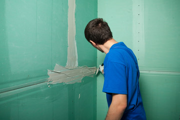 Portrait of young pretty man on construction site: male worker plastering walls with spatula while remodeling office