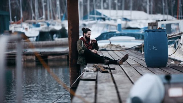  Rugged hipster man sitting alone on jetty in marina & playing harmonica
