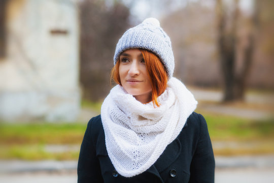 Portrait of a beautiful smiling woman with red hair in a big knitted white bactus scarf, a gray hat with a pompon made of natural wool and a black classic coat on a city street in the autumn day