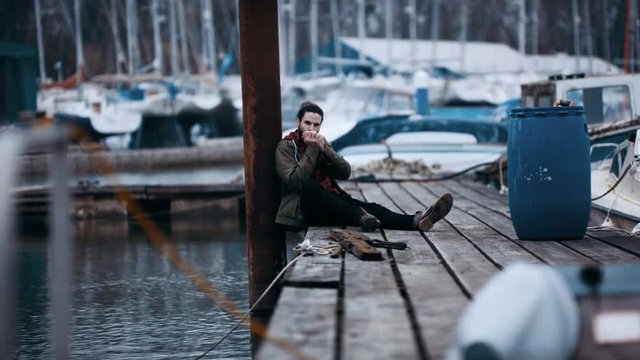  Rugged hipster man sitting alone on jetty in marina & playing harmonica