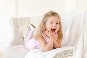 A little girl is lying on her stomach on the couch and laughing. Happy little girl on white sofa