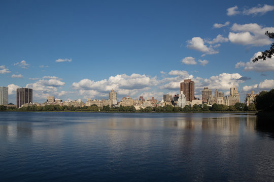 Urban city landscape of buildings and skyscrapers behind the lake or river and trees, with reflection of buildings in water and clouds on blue sky