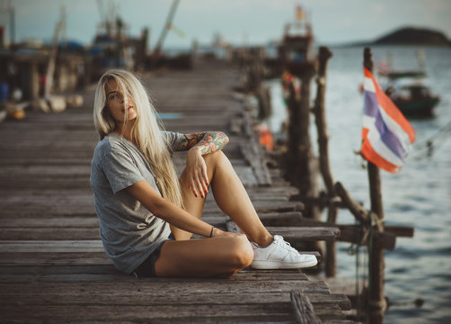Hipster girl sitting on wooden pier in the rays of the setting sun. A vacation in Thailand. Hands with tattoos