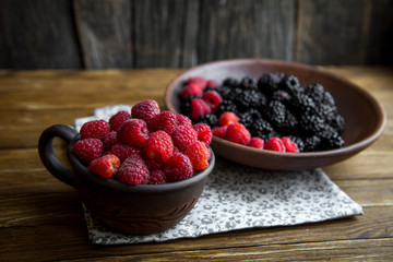 a ceramic mug with raspberries and a bowl with blackberries stand on a wooden table.