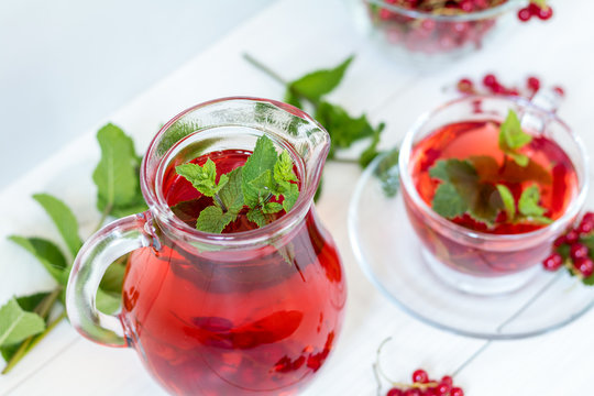 Redcurrant drink in transparent glass carafe and cup. Clear glass vase with red currant berries on the white wooden background