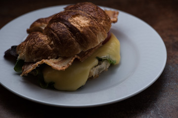 Croissant con quesa fundido, bacon y vegetales servido en plato blanco 
