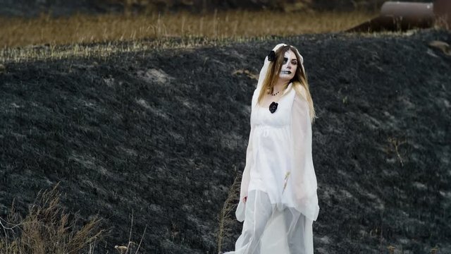 The young woman with spooky make-up of dead bride for Halloween dressed in a white wedding gown going through the field with burnt grass. 4K