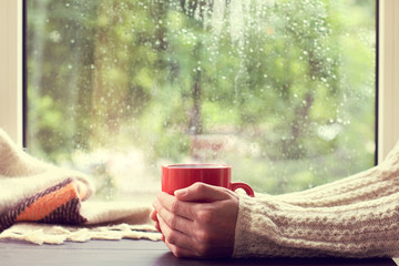 cozy home atmosphere in autumn/ red mug of hot drink in hand, when behind is window is rain