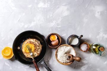 Homemade pancakes with fried orange in cast-iron pan and ingredients above. Wooden bowls of flour, yolk, salt, milk, olive oil over gray texture background. Top view with space. Home cooking concept
