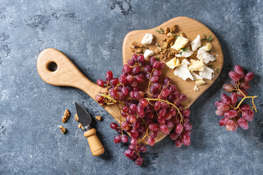 Wooden serving board with fresh red grapes, walnuts, goat and cheddar cheese, with cheese knife over blue texture background. Top view, copy space.