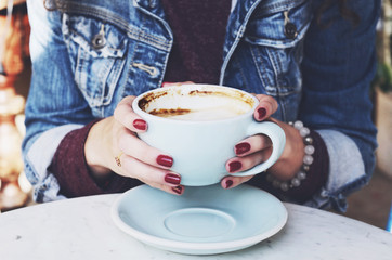 Woman in jeans jacket holding cup of cappuccino in her hands with red nails