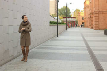 Young woman in a beige coat stands near a brick wall, does not look at the camera