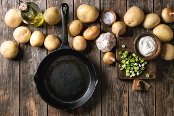 Obraz na płótnie Canvas Ingredients for cooking dinner. Raw whole washed organic potatoes, onion, garlic, salt, olive oil, cast-iron pan, spring onion and cream-fresh over old wooden plank background. Top view with space