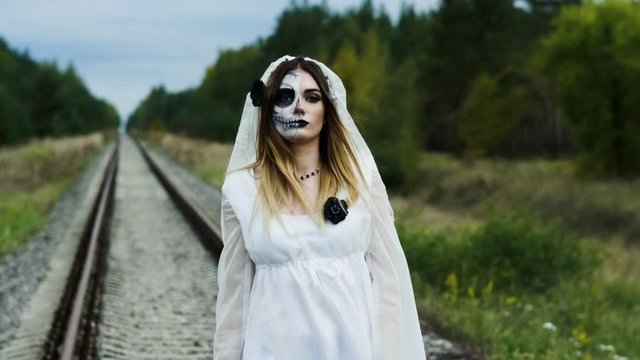 The young woman with spooky make-up of dead bride for Halloween dressed in white wedding gown is walking on the rails among the autumn forest. 4K