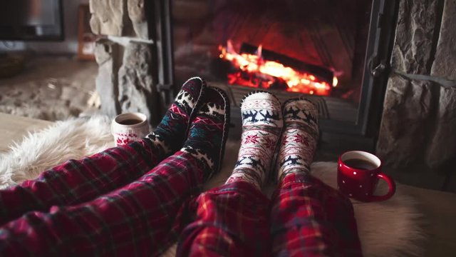 Feet in woollen socks by the Burning Christmas Cozy Fireplace. 4K. Couple relaxes by warm fire with cup of hot drink and warming up their feet. Winter and Christmas holidays concept.