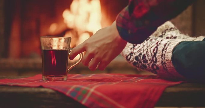 Woman relaxes by warm fire in woollen socks and having a cup of hot tea. Close up on feet. Cozy evening by the fireplace during the cold season. Slow motion 120 fps. Dolly shot. 4k graded from RAW.