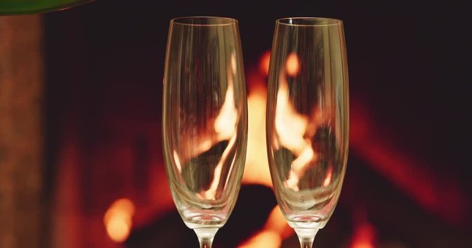 Pouring two glasses of sparkling champagne in front of warm fireplace. Slow motion. Zoom out. Dolly shot. 4k graded from RAW. 