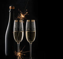 A bottle of champagne and two filled glasses on a black background, in one glass a Bengal fire burns.