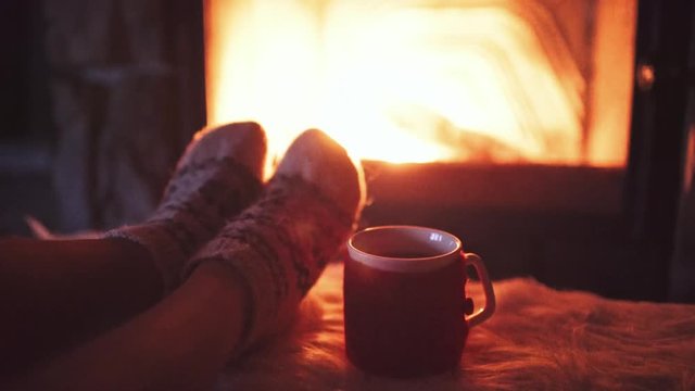 Feet in woollen socks by Burning Christmas Fireplace. 4K STABILIZED shot. Woman relaxes by warm fire with a cup of hot drink and warming up her feet. Close up. Winter and Christmas holidays concept.