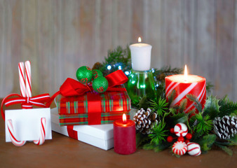 A Christmas setting of cheerful peppermint striped candle with presents, candies, artificial greenery and pinecones with a wooden background. Shallow depth of field used.