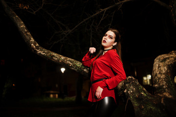 Black girl in red shirt and bright make-up on night street sitting on scary tree outdoor. Halloween theme.