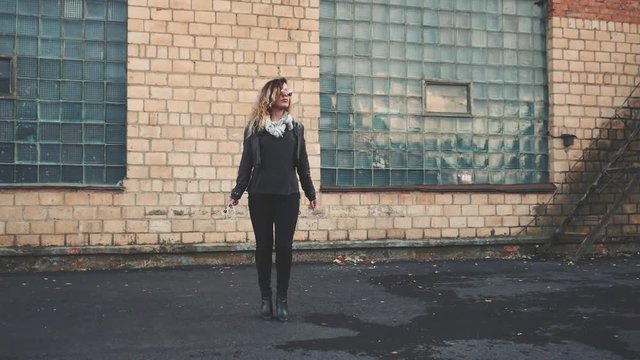 Young beautiful sexy woman with headphones and smartphone in hand in mirrored sunglasses, a black leather jacket, black jeans dancing by the wall of the industrial building of brick on the street.