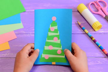 Child holds a Christmas greeting card in his hands. Child made a paper greeting card with a Christmas tree. Stationery on a table. Creating simple gifts crafts at home, at school or in kindergarten