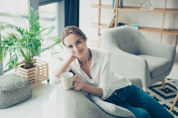 Cute brown haired gorgeous freelancer on the beige couch at home having hot beverage, looking in camera, so comfortable, in smart fashioanble outfit, with fashionable hairdo