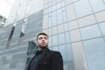 A young businessman with a beard and a black suit stands on the background of modern light architecture and looks up and down. Business portrait.