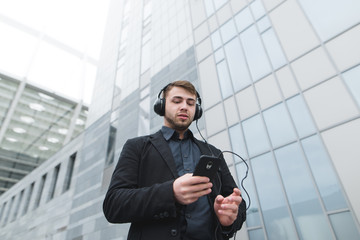 Fototapeta na wymiar A young business man listens to music in headphones and uses a smartphone while walking around the city. Against the background of modern architecture.