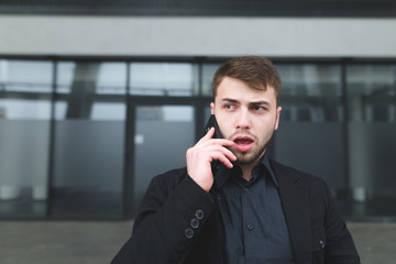 A street portrait of a young serious man with a beard that communicates on a mobile phone against a dark wall of modern architecture. Business Portrait of a Beautiful Hipster Businessman.